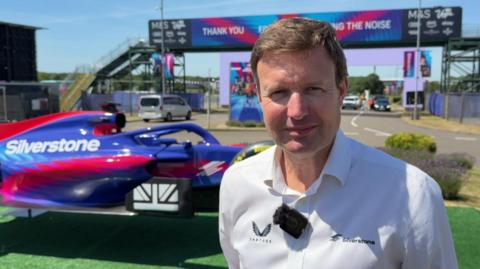 A head and shoulders shot of Stuart Pringle, outside at Silverstone. He has short, light brown hair and is wearing a white open-neck shirt, with a small black microphone attached to it. Behind him is a purple and red car with Silverstone written on it in white letters and beyond that a road with a bridge over it.