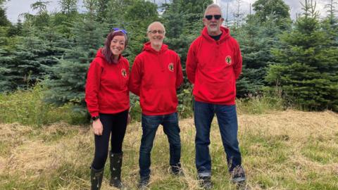 Organisers Hannah Johnson, Scott Crowther and Paul Flower standing in a field in front of Christmas trees, wearing red hooded tops