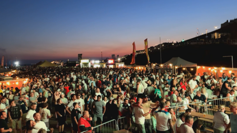 Hundreds of football fans fill a seafront fan park in Brighton at dusk, with food stalls, tents and large screens visible beneath a clear evening sky.