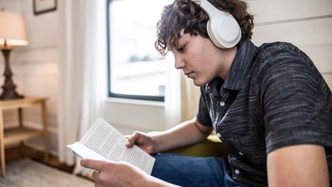 GCSE English language student wearing headphones at home with book listening to Bitesize podcast