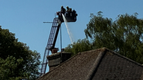 Firefighters operate an aerial ladder platform above a building roof, directing a jet of water downwards. The extended ladder rises above nearby trees, illustrating the specialist equipment used to tackle fires from height.