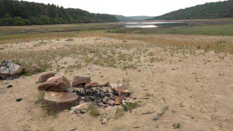 a sandy area with a few patches of green grass. Int he foreground there are a few rocks, and in the background, the remains of a reservoir can be seen. There are green hills with trees on both sides.