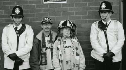 A black and white image of a male and female football fan wearing hats, badges, scarfs and football shirts. He has his arm around her. The are standing between two West Midlands Police officers who have helmets, and uniform on. They are both wearing black gloves and have their hands crossed in front of them. 
