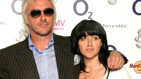 A man with platinum blond hair with sunglasses and wearing a black suit and light blue shirt, standing next to a woman with black hair and a white shirt.