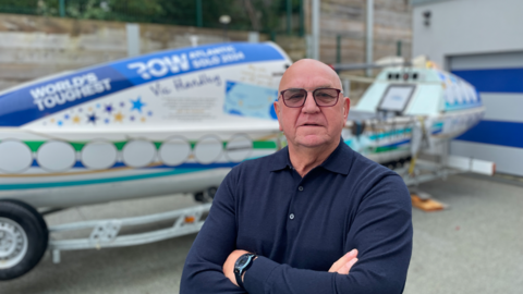 A bald man with glasses, wearing a navy blue shirt, stood in front of a white boat