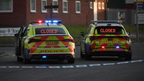 Two police cars marked yellow, orange and blue with flashing blue and red lights parked on a road at a junction facing away from the camera. There is a blue and white police cordon between the cars and the camera.