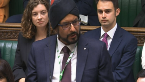 Warinder Juss, wearing a dark suit, stands in the House of Commons, flanked by other MPs sitting behind him