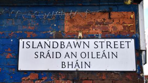 A white bilingual street sign in Irish and English. It is on a red brick wall that is partially painted blue. Some barbed wire can be seen on the wall above the sign. 