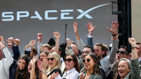 Group of people in the foreground raising their arms and clapping in front of a large stage backdrop displaying the SpaceX logo.