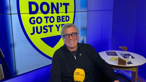 A man with grey and thick black-rimmed spectacles sits in a blue-lit recording studio. A blue and yellow badge with the words 'Don't go to bed just yet' is on the wall behind him. A BBC Radio Leeds mic is in front of him.