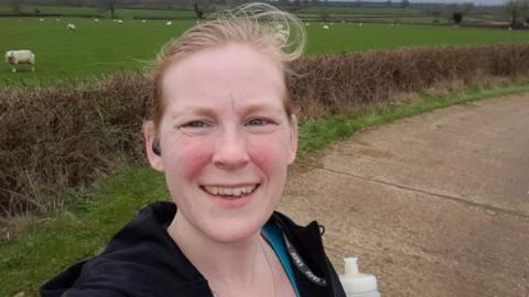 A selfie of Toni Bull taken while on a run. She is standing on a concrete road with a hedge and fields in the background.