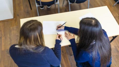 Two girls with long brown hair sit at a school desk writing on a piece of paper. 