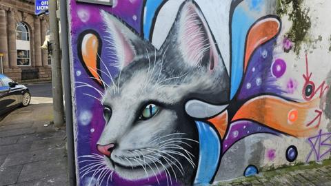 A grey cat with blue and green eyes looking to the left amongst swirls of orange and purple on one of the street murals