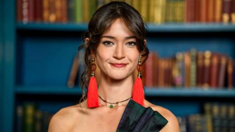 A head and shoulders photo of a young woman with dark hair tied back, with loose curls around her face. She is wearing red earrings with long tassels, a green jewelled choker necklace and the top of a tartan bow on a shoulderless corset can just be seen. Behind her are blue painted shelves with books.