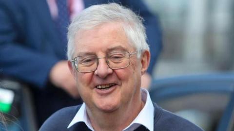 Mark Drakeford smiling to the camera, wearing a white shirt and blue jumper.