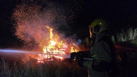 A firefighter in full protective gear including a yellow helmet, is holding a large hose reel jet which is spraying water. In the background is a large fire with wood in it, and orange flames and sparks coming off it 