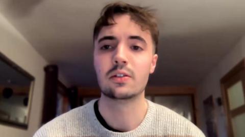 Eòin Cuimeanach na shuidhe air beulaibh camera agus geansaidh soilleir air.