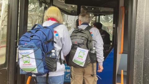 Nick with short white hair and Kerstin with short blonde hair wearing grey sweatshirts and backpacks with Harry's Pals leaflets attached. They are seen from the back entering a bus via folding electric doors.