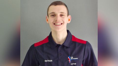 A teenage boy, wearing a GB deaf swimming polo shirt, smiles at the camera.