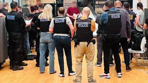 Police officers, some in uniform and some plain-clothed, all wearing stab vests, are briefed in a room with a wooden floor.