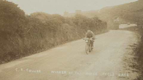 A sepia image from 1907 of the first TT race - a man is on an early motorbike heading from right to left down a road lined with a verge.