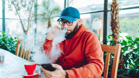 Man in red hoody and blue baseball cap blowing a cloud from a vape while looking at his phone with a teacup in front of him