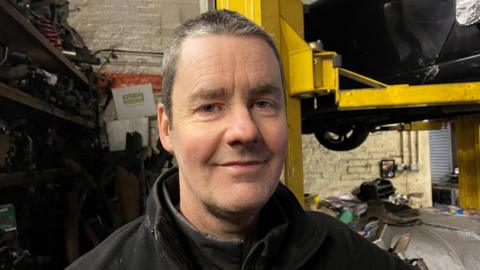 Paul Jones is a mechanic standing in his garage in front of a car which has been raised up on a car lift. There is a stack of car parts behind him. He is looking at the camera and smiling. He has short grey hair and is wearing black overalls.