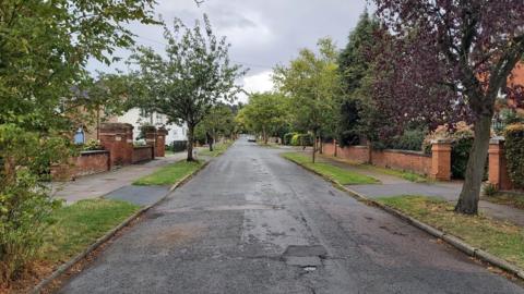 Eldon Road, Cheltenham. It is a wide residential street with a pavement on either side, which is lined with trees and grass. There are low brick walls at the end of everyone's front gardens, and no cars are parked on the street.