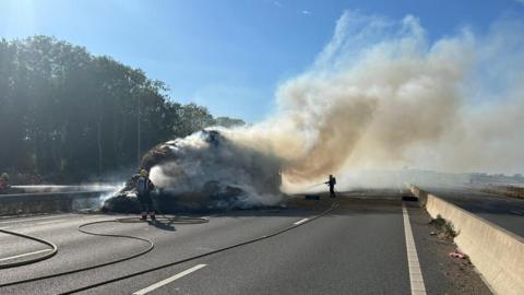 An empty dual carriageway with a large object engulfed in fire. Several firefighters stand nearby pointing hoses towards the fire. The sky is blue and there are trees on the left. The right carriageway is empty.