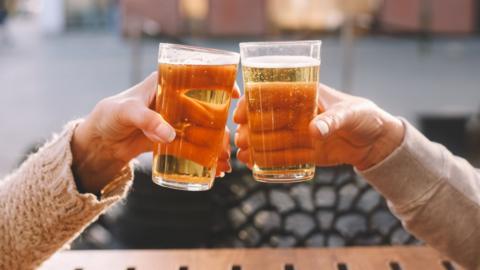 Close-up of two people toasting with pint glasses of beer while sitting at an outdoor wooden table. 