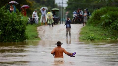 A shirtless man wearing a brown hat and holding his flip flops in one hand wades across waist-deep water while people watch from the other side of an inundated road in El Progreso, Honduras.