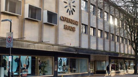 A photo showing the outside of the Cavendish House building in Cheltenham, with a clock and sign and windows below
