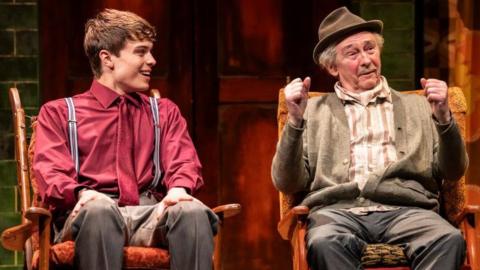 Tom Major in a red shirt with a red tie and grey trousers. He is sat next to Paul Whitehouse who is dressed in grey trousers, a white and brown striped shirt and cardigan. He is wearing a hat. Both men are looking away from the camera. 