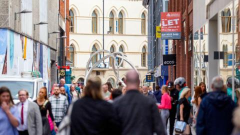 A man and woman walk in front of a busy Belfast street. Al that are in the foreground are out-of-focus. In the background tall cream building and a round iron structure can be seen.