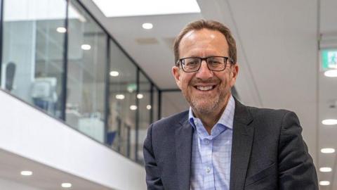 Laurence Frewin photographed at the college. He is seen wearing a grey suit with a light blue shirt. He has stubble and brown swept back hair. He is also wearing glasses and smiling at the camera.