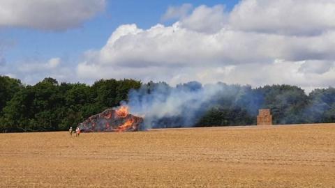 A burning haystack in a field with smoke rising from it. There is a second undamaged haystack in the background