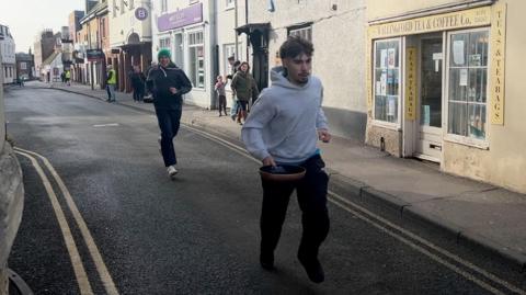 Two men run down a narrow road between shops in the town holding frying pans.