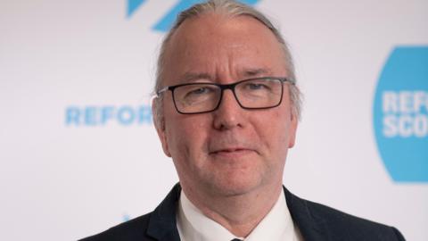 David Kirkwood - man with grey hair and black rimmed glasses, wearing a black suit and white shirt, standing in front of a Reform backdrop