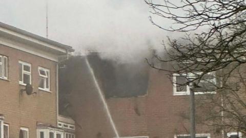 A close up of a flat with a gaping hole in the roof and smoke emitting from the hole. A stream of water is pointed at the hole
