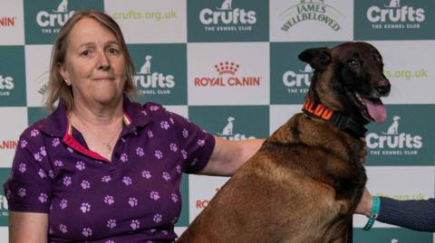 Owner Mandy with thin dark blonde hair, wearing a purple short sleeve polo with paw prints on it next to Baloo