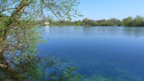 A large lake surrounded by green trees. There is a small white building on the left bank in the distance