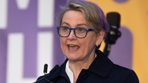 Home Secretary Yvette Cooper speaks on stage wearing bright blue-rimmed spectacles. She has short, cropped, dark blonde hair and wears small silver earrings.