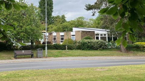 A brick one-storey building with windows and surrounded by trees. There is a road, a pavement, a bench, a waste bin and a lamp post in front.