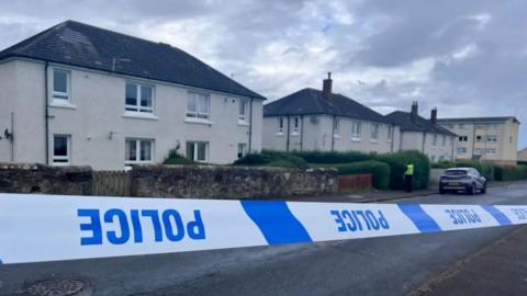 Blue and white police tape which is upside down in front of a row of cream two-storey buildings. A police officer is visible in a distance.