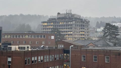 A view of Wycombe Hospital building on an overcast day, with lights visible in the windows and trees and other buildings in the background.