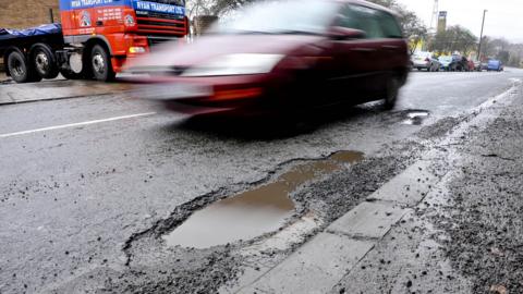 A car drives past a pothole on a road. Other vehicles can be seen on the road. 