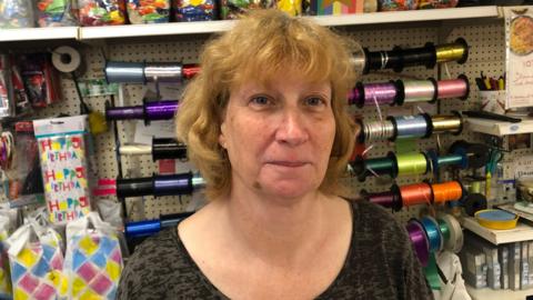 Looking at the camera with a closed lip smile is Julie Lambert who has strawberry blonde wavy hair and a fringe. She is standing in front of different coloured ribbons and birthday banners which are on reels in her "Greetings" card and gift shop in March. She has freckles across her nose and cheeks and a birth mark on her jaw. 