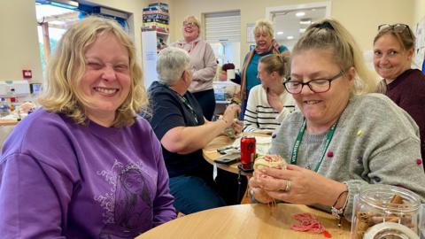 Five women sit at two round tables laughing and two of them are also seeing. Two other women can be seen standing in the background, one speaking and the other also laughing. The lady closest on the left is wearing in a purple jumper with short blond hair. The lady closest on the right is wearing a grey jumper with small colourful dots. She is one of the ladies sewing. They are all inside the room of a community centres with cream walls and an open door leading to the outside.