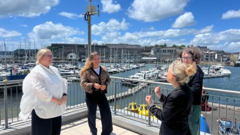 on the left and stood next to Rebecca Hegedus. They are stood opposite their interpreters. They are all smiling at one another and are stood on the balcony with a view of Queen's Anne Battery where there are docked boats. 