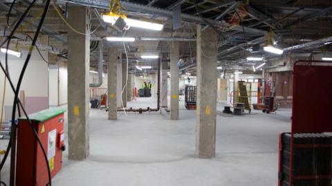 Image of the inside of a building under construction. The building has an open ceiling showing all the ductwork, a concrete floor, a number of concrete pillars and strip lighting. In the foreground is a red metal power station. 